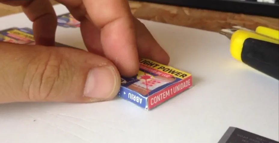 Mock-up físico da embalagem montado e segurado entre dois dedos, ao lado das ferramentas de corte usadas para montá-lo: o conceito tangível com os próprios recursos do instituto.